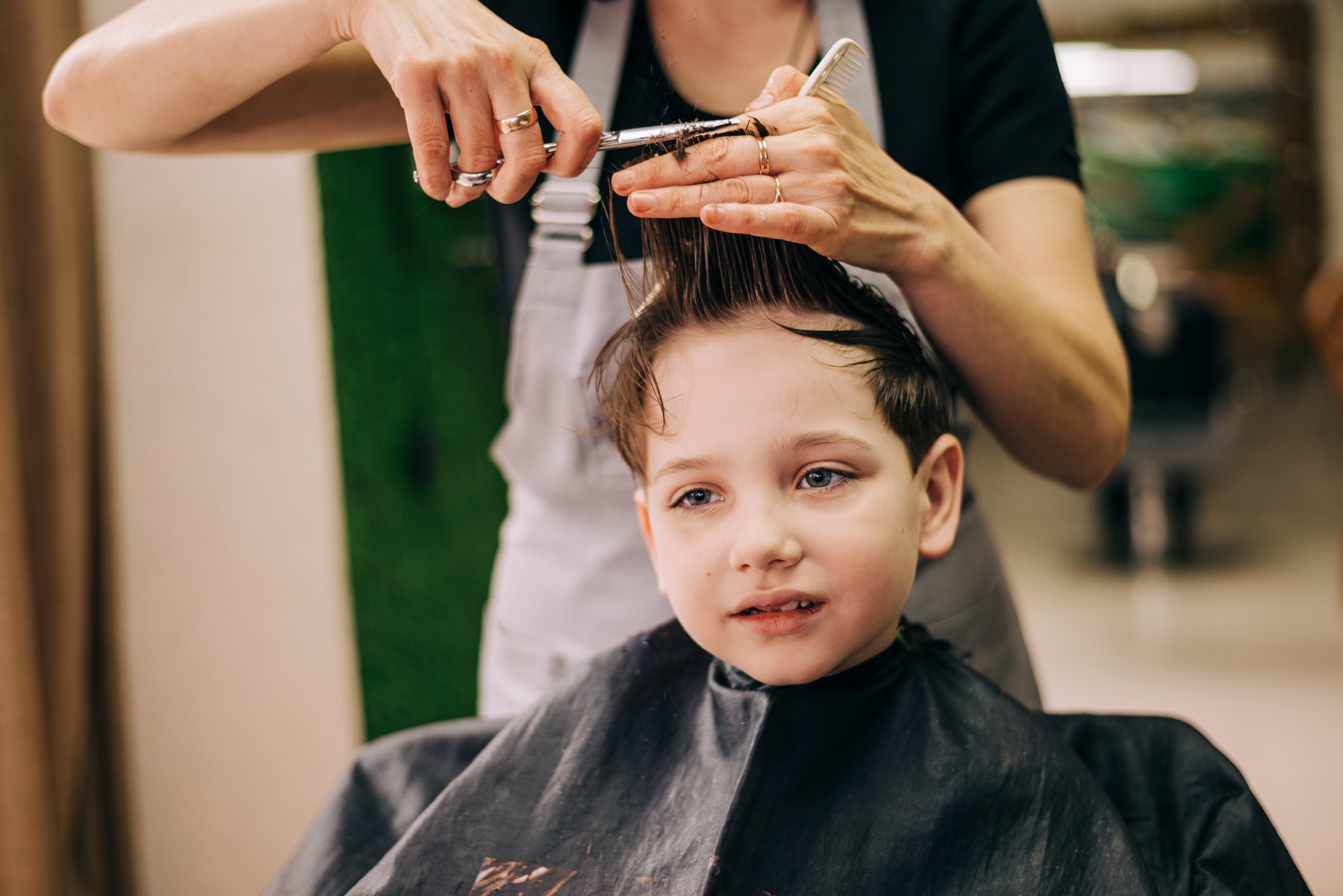 Little kid boy in the barbershop or hairdressing saloon. Barber is doing haircut for a toddler